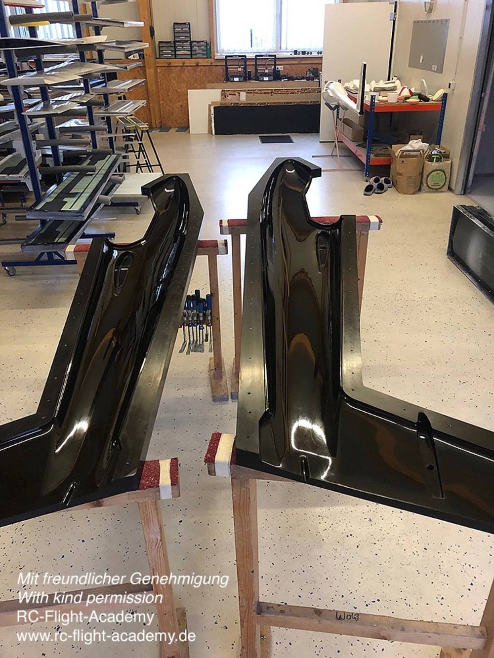 Zwei glänzende, schwarze Teile auf Holzgestellen in einer Werkstatt mit Regalen und Materialien im Hintergrund.