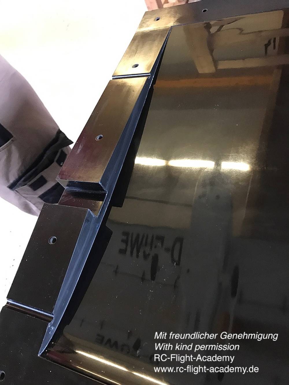 Nahaufnahme eines glänzenden Metallblechs mit einem abgeschrägten Rand und sichtbaren Schraubenlöchern. Text auf dem Blech: 