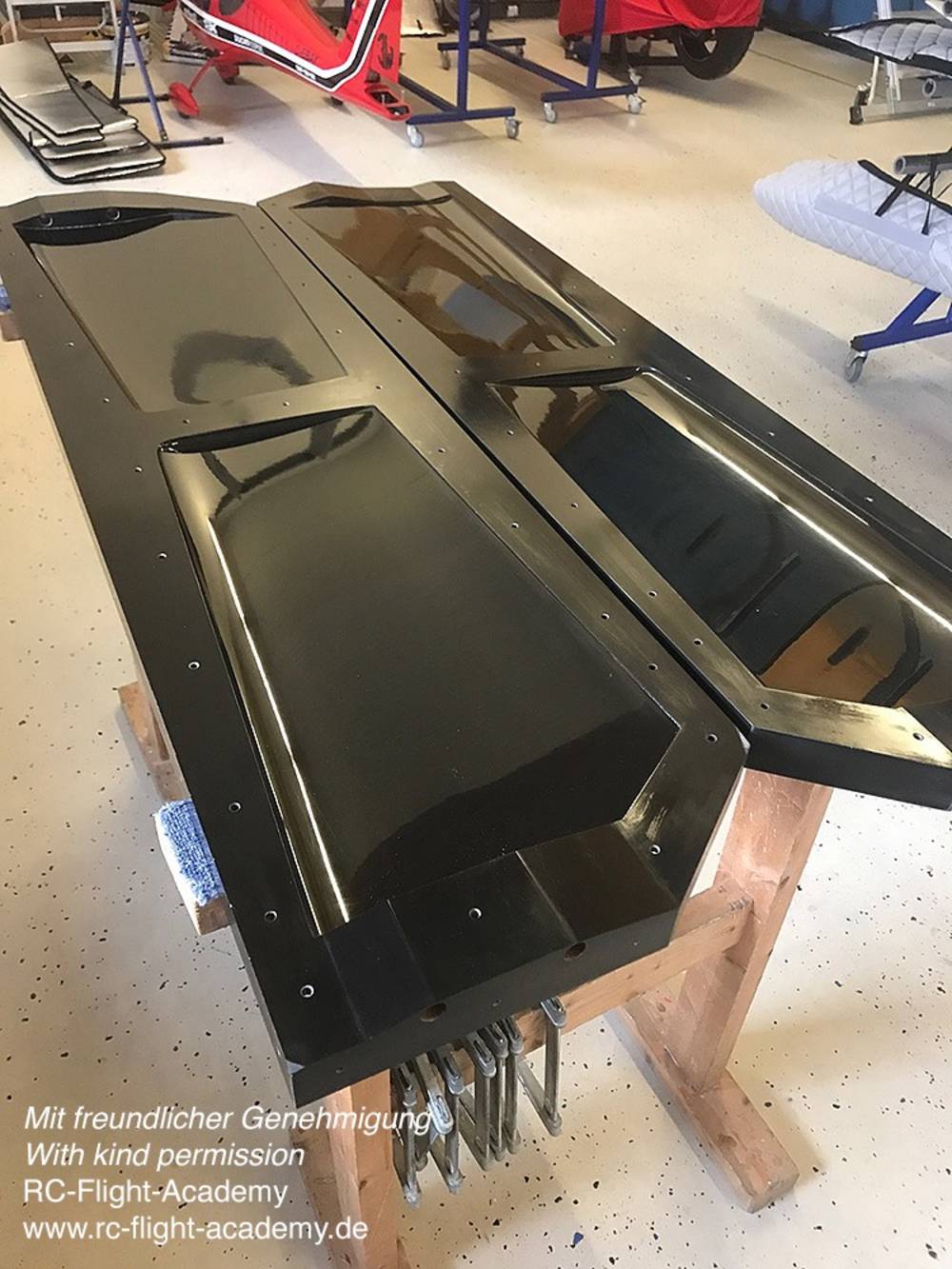 Schwarze Flugzeugteile auf einem Holztisch in einer Werkstatt. Im Hintergrund ein Modellflugzeug und weitere Werkzeuge.