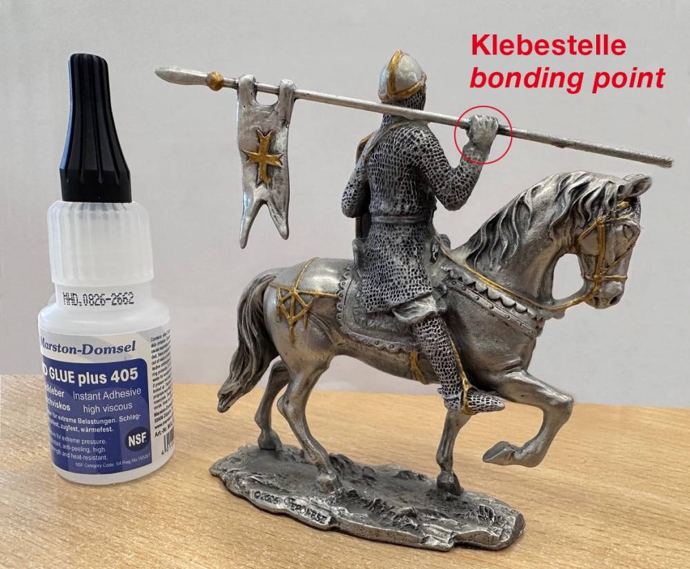 Eine Ritterfigur auf einem Pferd mit einer Lanze, daneben eine Tube Kleber. Der Klebepunkt ist markiert.