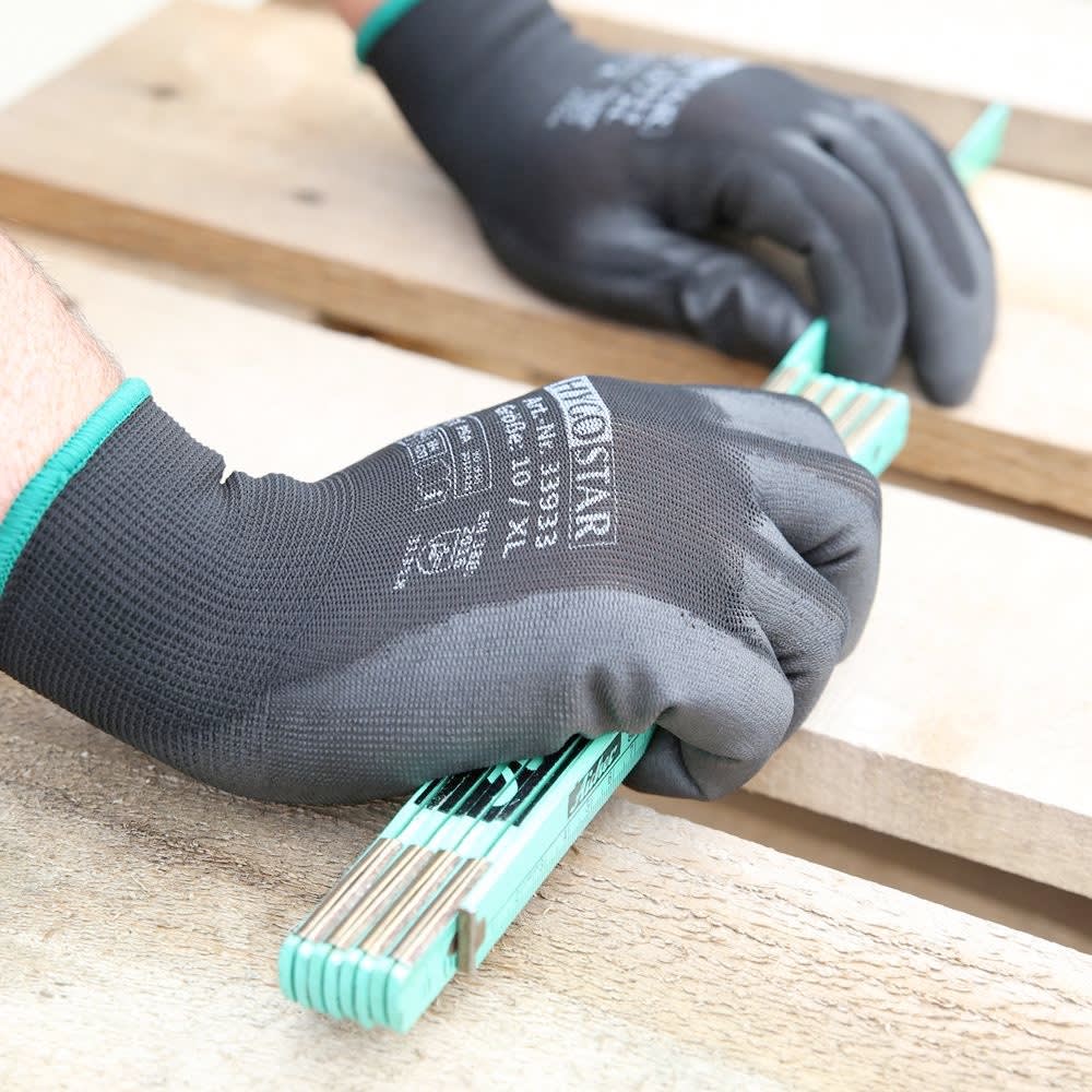 Zwei Hände in schwarzen Handschuhen halten einen grünen Zollstock auf einem Holztisch.