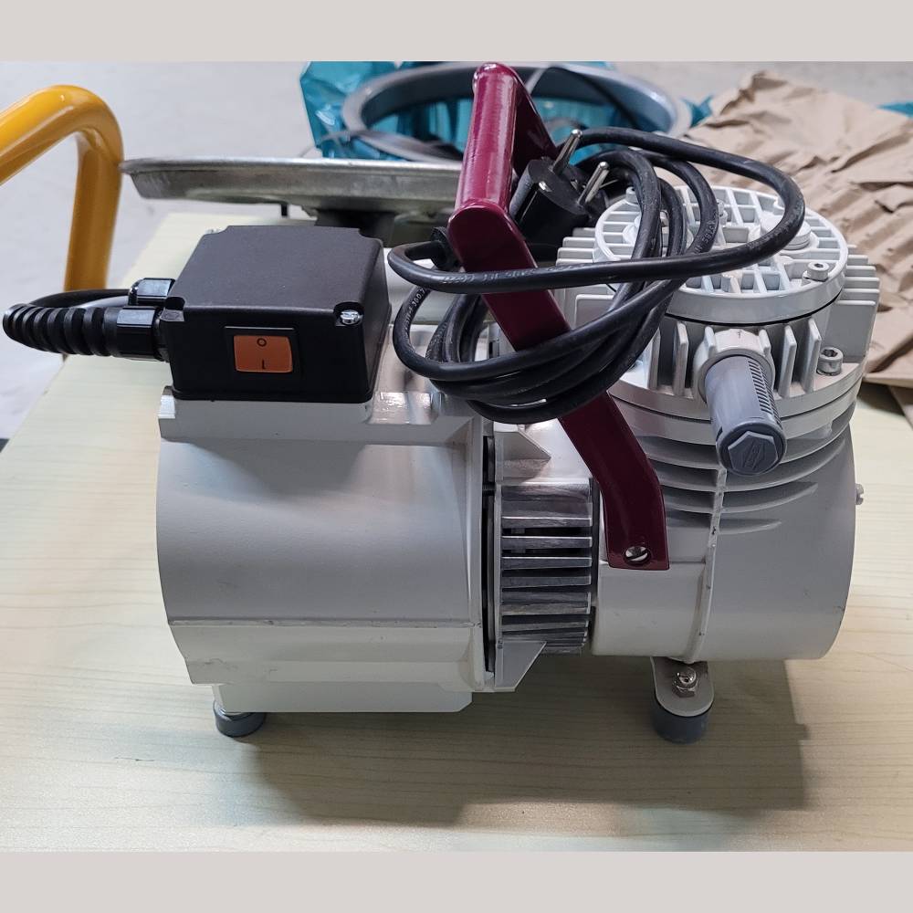 An electric vacuum pump on a table with an on/off switch and a cable. The pump is gray with a red handle.