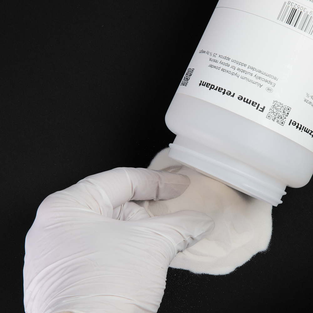 Eine behandschuhte Hand hält ein weißes Pulver, das aus einem weißen Behälter auf eine schwarze Oberfläche geschüttet wird. Der Behälter hat ein Etikett mit Text und QR-Codes.