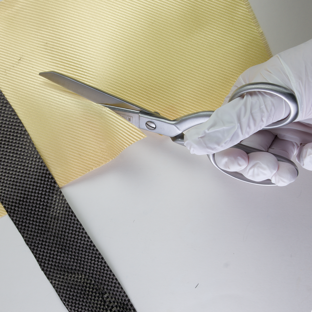 Eine Hand in einem weißen Handschuh schneidet mit einer Schere ein gelbes Material, daneben liegt ein schwarzes Material mit Karomuster.