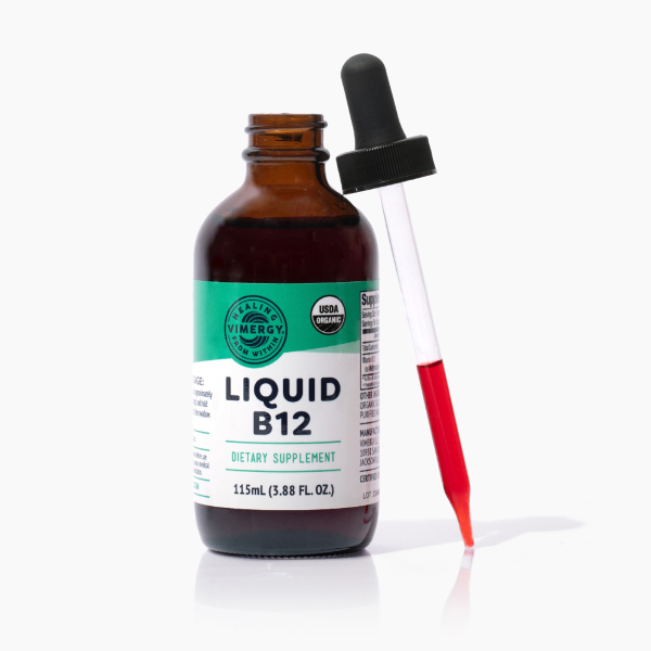Produktfoto: Eine braune Glasflasche mit einem Etikett, auf dem 'Liquid B12' steht. Daneben liegt eine Pipette mit roter Flüssigkeit. Die Flasche ist als USDA Organic gekennzeichnet und enthält 115ml Nahrungsergänzungsmittel.